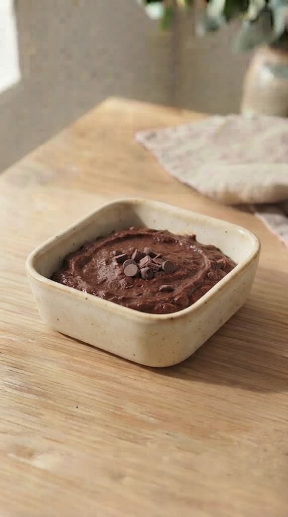 Dark egg-free brownie batter smoothed into a small square cream ceramic baking dish with a few reserved chocolate pieces on top