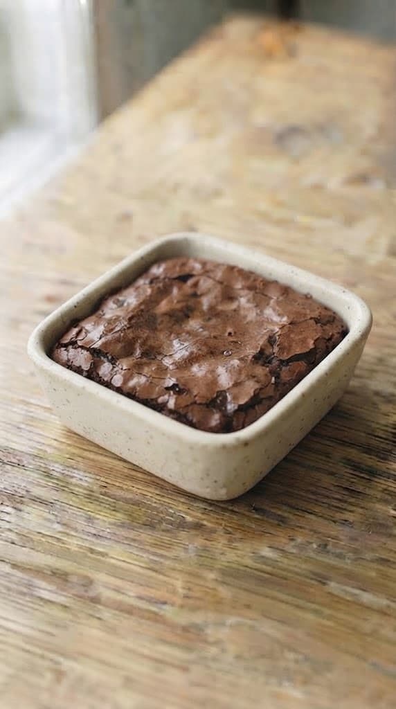 Freshly baked fudgy brownie in a small square ceramic baking dish with a thin crackly top and soft dark center