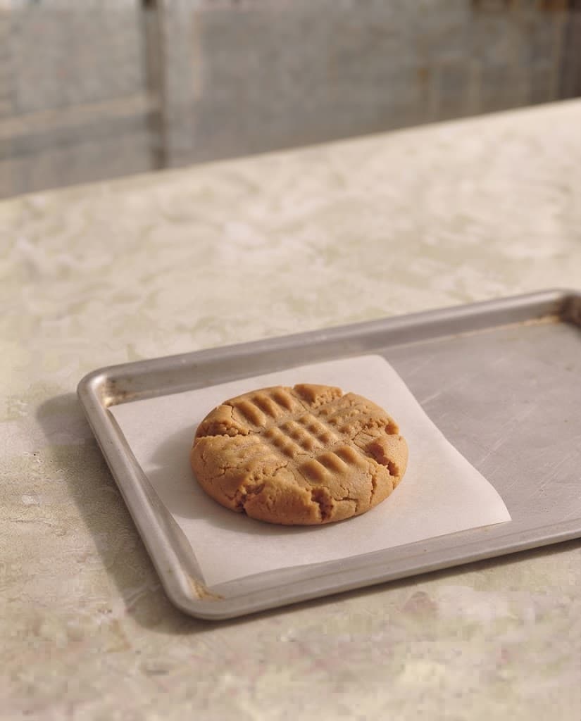 Freshly baked peanut butter cookie with lightly golden edges and visible fork marks resting on a parchment-lined baking tray